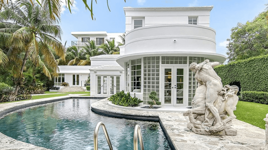 A large, two story Art Deco Home with a pool that curves around the back entrance flanked in a tropical theme with large palms and a Greek Sculpture depicting a horse and man.