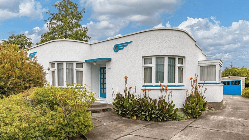 A one story Art Deco home, in white with blue trim and a paved front framed with foliage, located at 1234 House Lane, Langsley, VA 90210 and listed for $650,000.