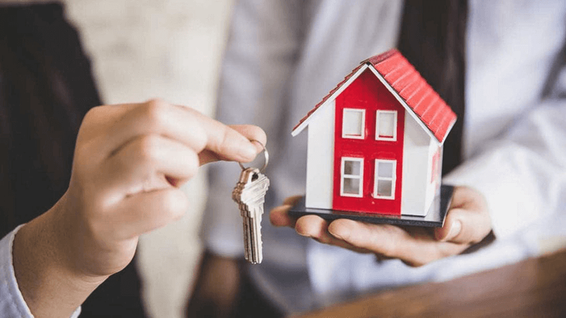 A hand holds a set of new keys while another hand holds a model of a red and white two story home.