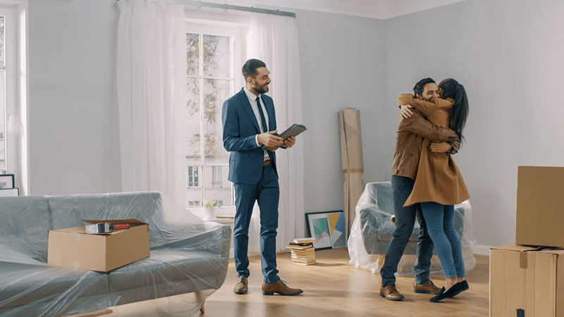 A realtor watches as a heterosexual couple celebrates their new home purchase by hugging in their packing box strewn living room.